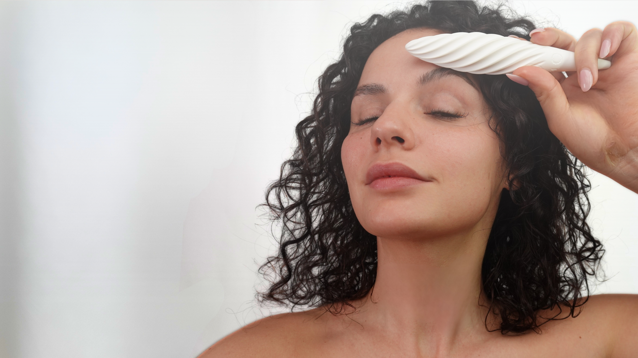 Young woman with curly hair using the white Fevowell Facer above her left eyebrow during a calm selfcare ritual