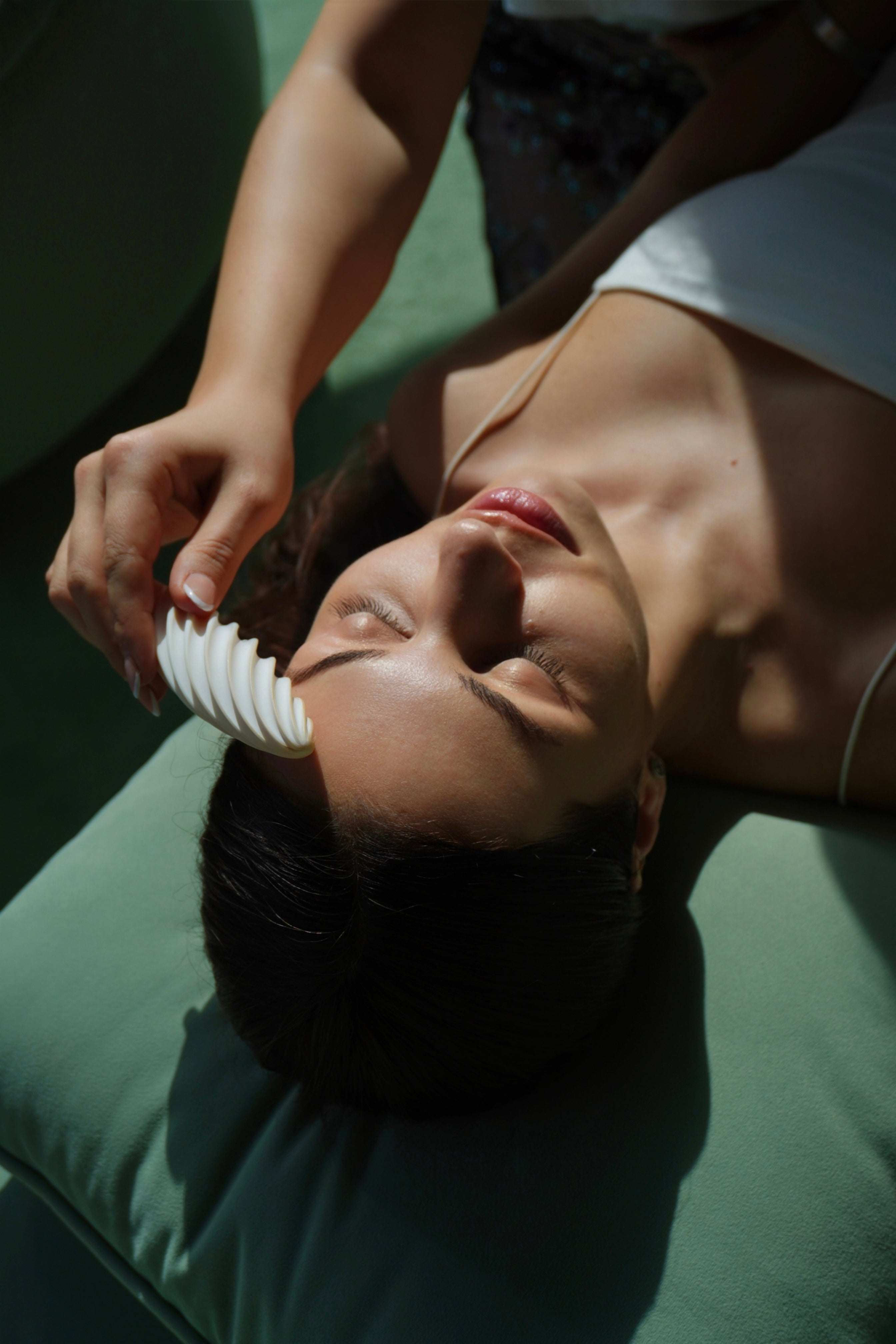 Woman lying down in natural sunlight, massaging her forehead with the Fevowell Facer for a calming and revitalizing selfcare ritual