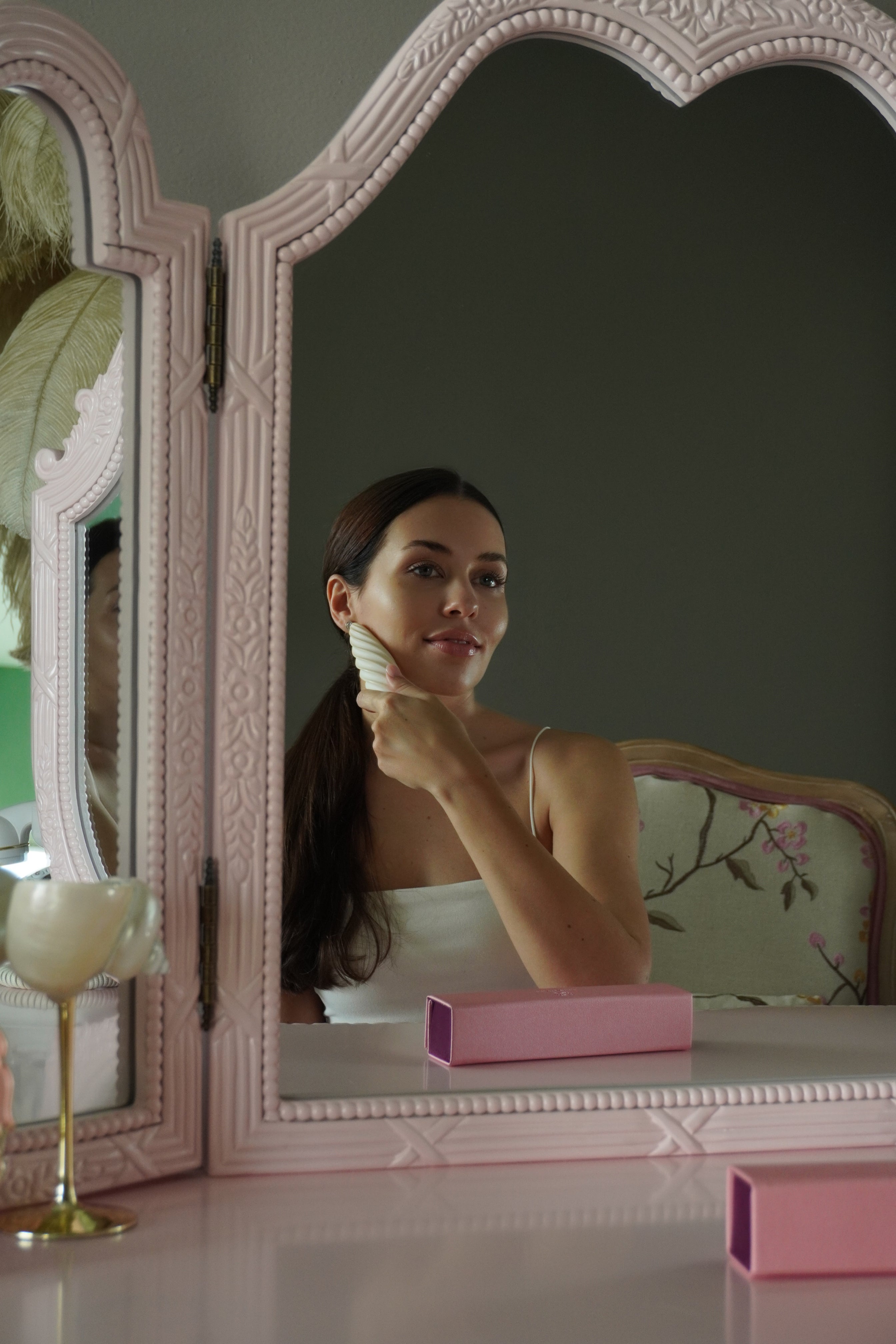 Woman using the Fevowell Facer in front of a vintage mirror, creating a calm beauty ritual with the pink product box in view